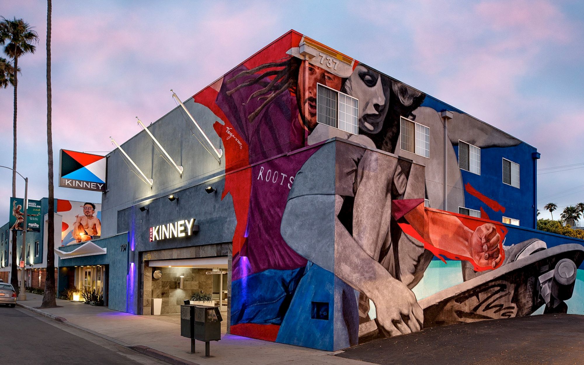 A vibrant mural covers a building exterior featuring artistic figures. The building is marked "KINNEY" and palm trees line the street.