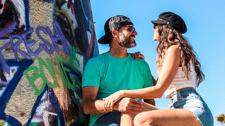 A couple sits on a graffiti-covered wall, smiling at each other while wearing casual clothes and hats against a clear blue sky.