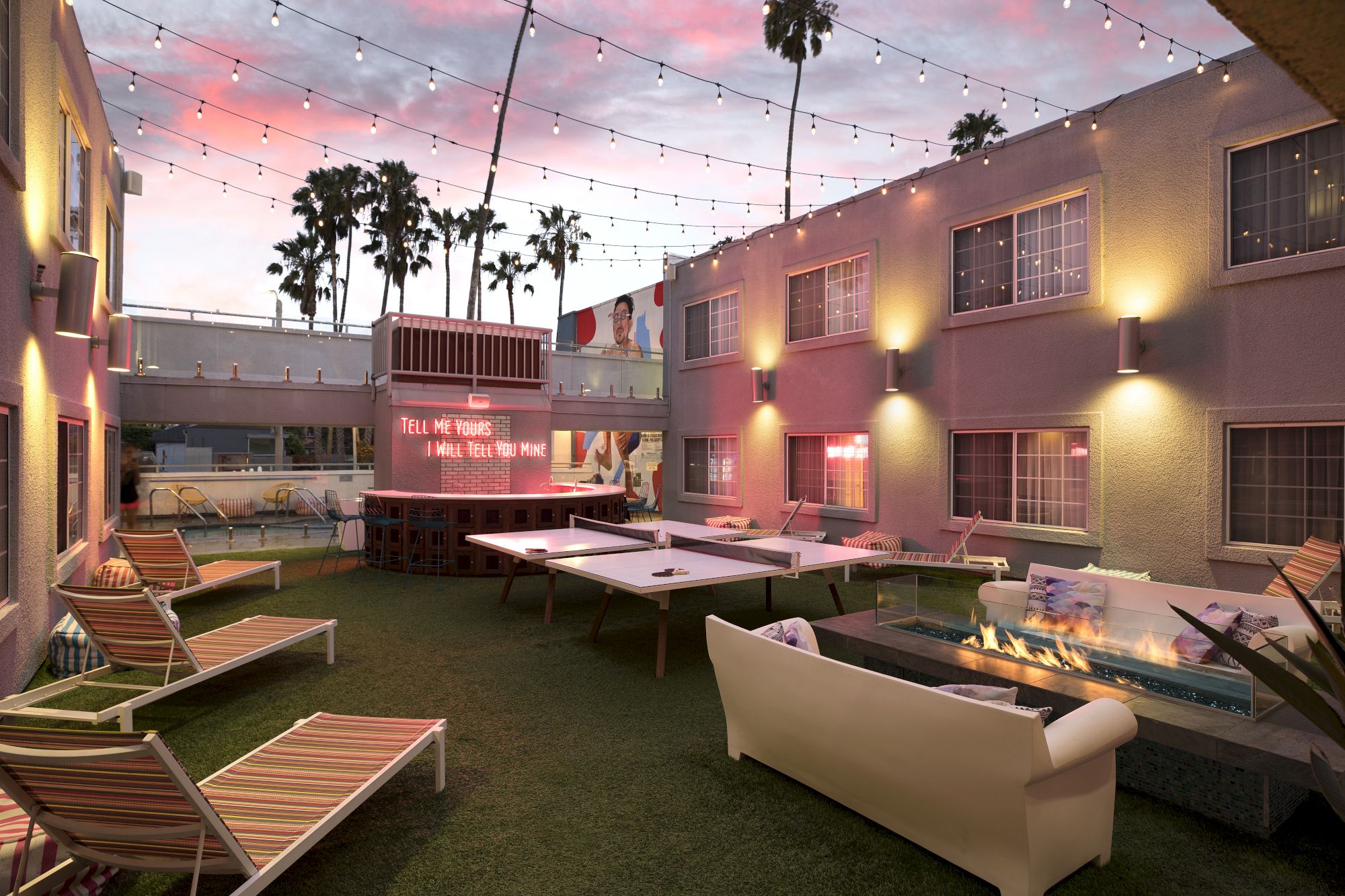 Outdoor patio at sunset, string lights, lounge chairs, ping pong, fire pit, and a neon sign reading "Tell Me Yours I Will Tell You Mine."