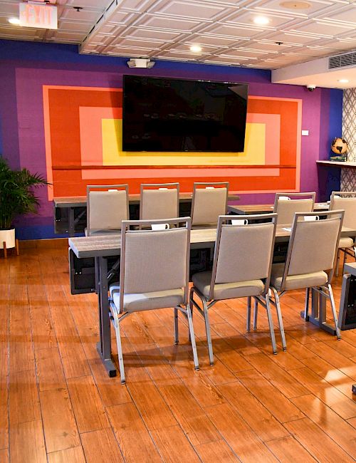 A modern conference room with chairs facing a large TV on a colorful wall, wooden floor, and a long side table along a patterned wall.
