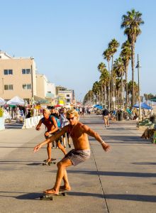People are skateboarding on a sunny boardwalk next to a beach, with palm trees and various stands around.