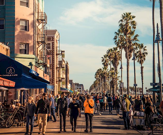 A bustling boardwalk lined with palm trees, people walking, and a café on the left under a clear blue sky.