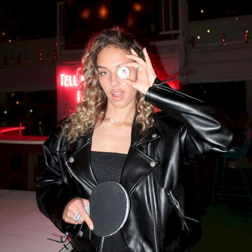A person in a black leather jacket holds a ping pong paddle and a ball, covering one eye, against a nightlife background.