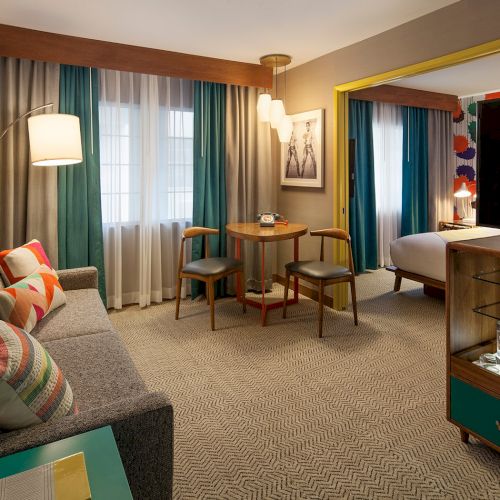 A modern hotel room with a sofa, TV, table, and large window. There's a bed in an adjacent room, visible through a wide doorway.
