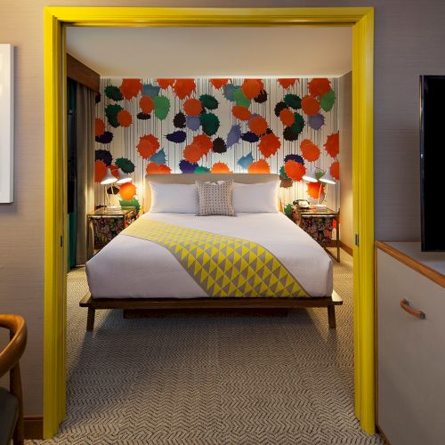 A colorful hotel room with a double bed, abstract wallpaper, a TV, minibar, and a framed artwork of a person on the wall.