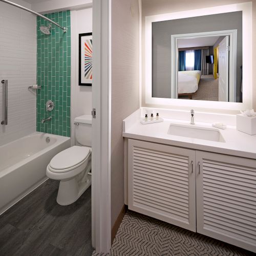 This image shows a bathroom with a white countertop, sink, toilet, bathtub, green-tiled wall, and a mirror reflecting another room.