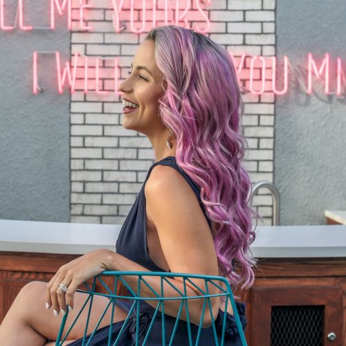 A person with pink hair is sitting on a chair, smiling, with neon signs saying "Tell me yours, I will you mine" in the background.
