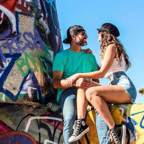 A couple sits on a graffiti-covered surface under a bright blue sky, smiling at each other. They are casually dressed in summer attire.