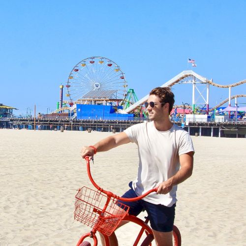 A person rides an orange bike by a beach, with a ferris wheel and roller coaster in the background on a sunny day.