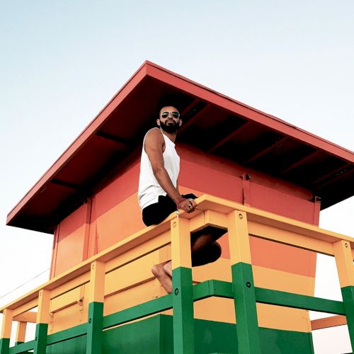 A person with sunglasses sitting on a colorful lifeguard tower with a clear sky background.