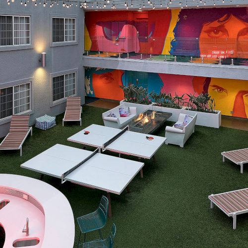 Outdoor courtyard with seating, lounge chairs, ping pong tables, vibrant wall art, and string lights overhead, creating a lively atmosphere.