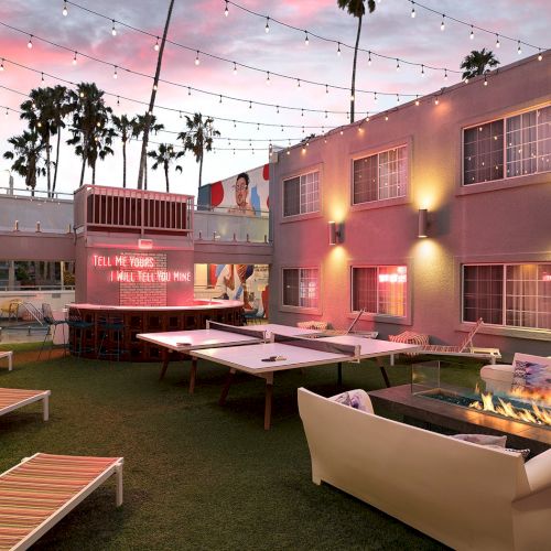Outdoor lounge with string lights, a fire pit, ping pong tables, lounge chairs, neon sign, and palm trees, creating a lively atmosphere.
