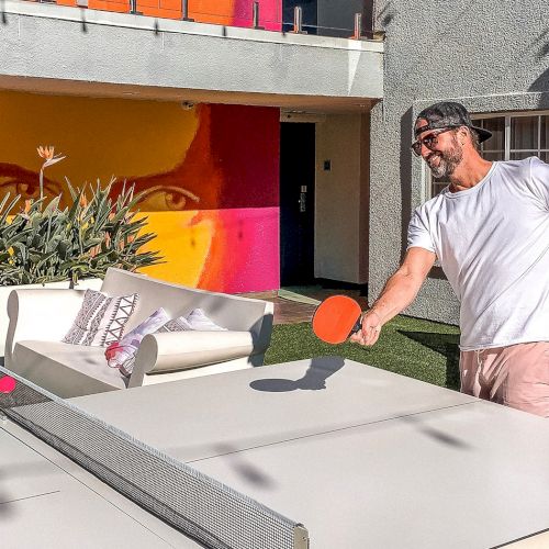A person playing table tennis outside, with colorful wall art and plants in the background.