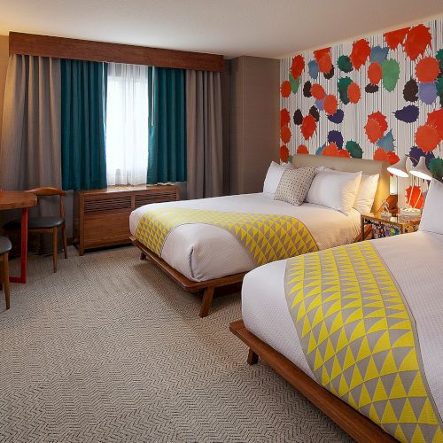 A hotel room with two beds, colorful wall art, yellow-patterned bed runners, a TV, and a small desk with a retro phone and chair.