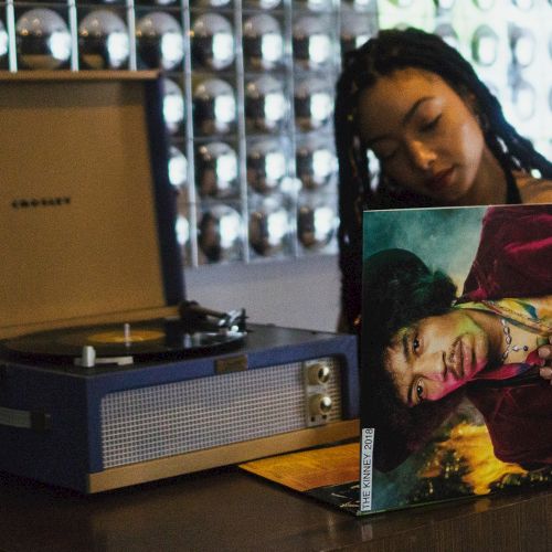 A person holds a Jimi Hendrix vinyl record near a Crosley turntable, with a wall of metallic spheres in the background.