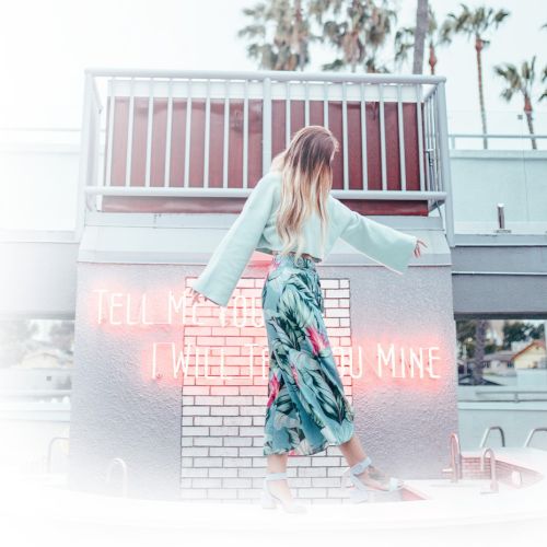 A person in a floral dress walks on a white surface near a pink neon sign reading "TELL ME YOU [ ] I WILL TELL YOU MINE."