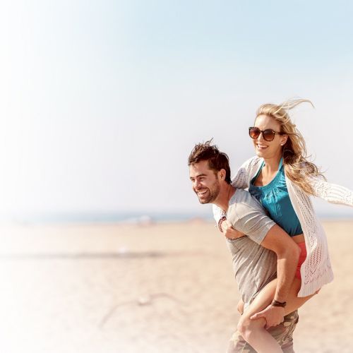 A man is giving a piggyback ride to a woman on a sandy beach. They both appear happy, with the woman wearing sunglasses and extending her arms.