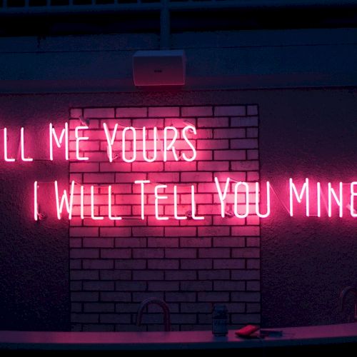 A neon sign says, "Tell me yours, I will tell you mine," on a brick wall in pink light.