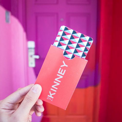 A hand holds a red envelope labeled "KINNEY" with patterned cards inside, in front of a vibrant pink and orange door.