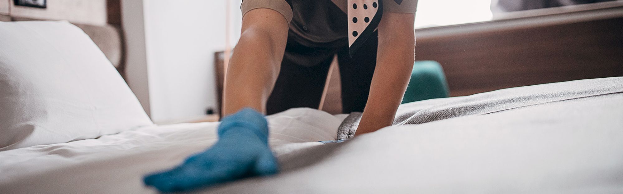 A person is making a bed, wearing a blue glove and a uniform, smoothing out the sheets in a room.