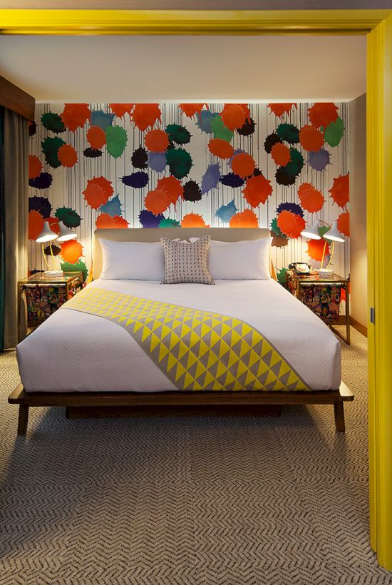 A colorful bedroom with a patterned wall, cozy bedding, a TV, and a well-lit space featuring modern decor and a beverage tray.