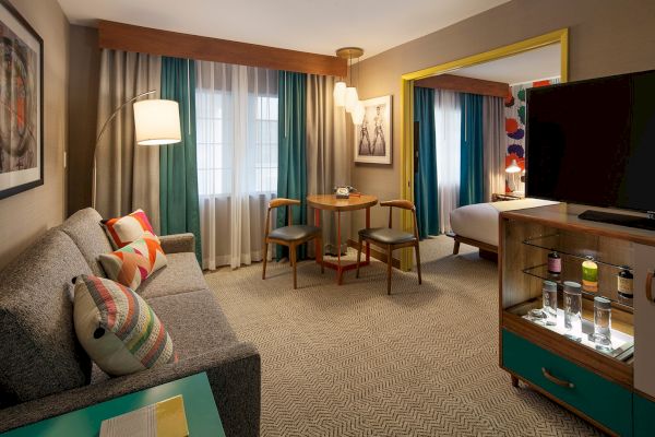 The image shows a modern hotel room with a sofa, table, chairs, TV, and bed. Bright accents and contemporary decor complete the space.