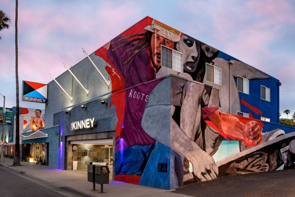 A building with vibrant mural art depicting people and abstract shapes, under a pink sky, labeled "KINNEY."