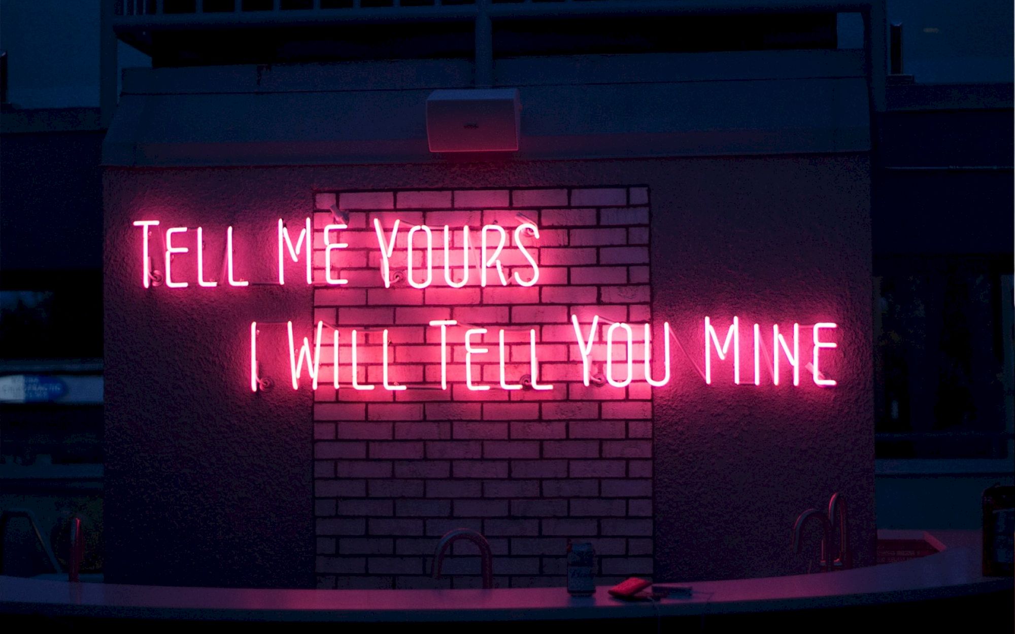 A neon pink sign reads, "TELL ME YOURS I WILL TELL YOU MINE," on a brick wall above a bar counter.