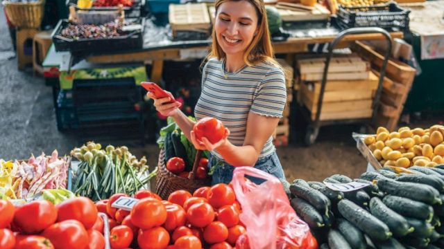 A person is choosing tomatoes at a market stall with various fresh vegetables, smiling and enjoying the shopping experience.
