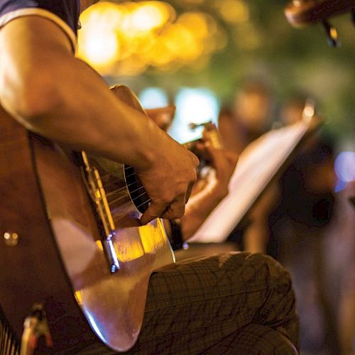 A person is playing an acoustic guitar, surrounded by warm, blurred lights in the background, creating a cozy atmosphere.