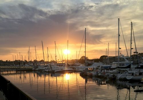 A marina with several sailboats docked at sunset, reflecting on the water, creates a serene and picturesque scene.