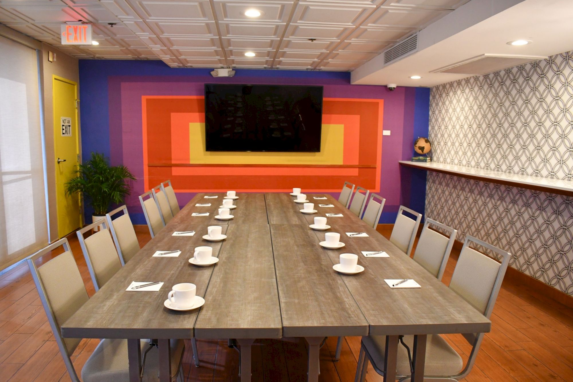 A conference room with a long table, set for a meeting with cups and napkins, and a TV on a colorful wall.