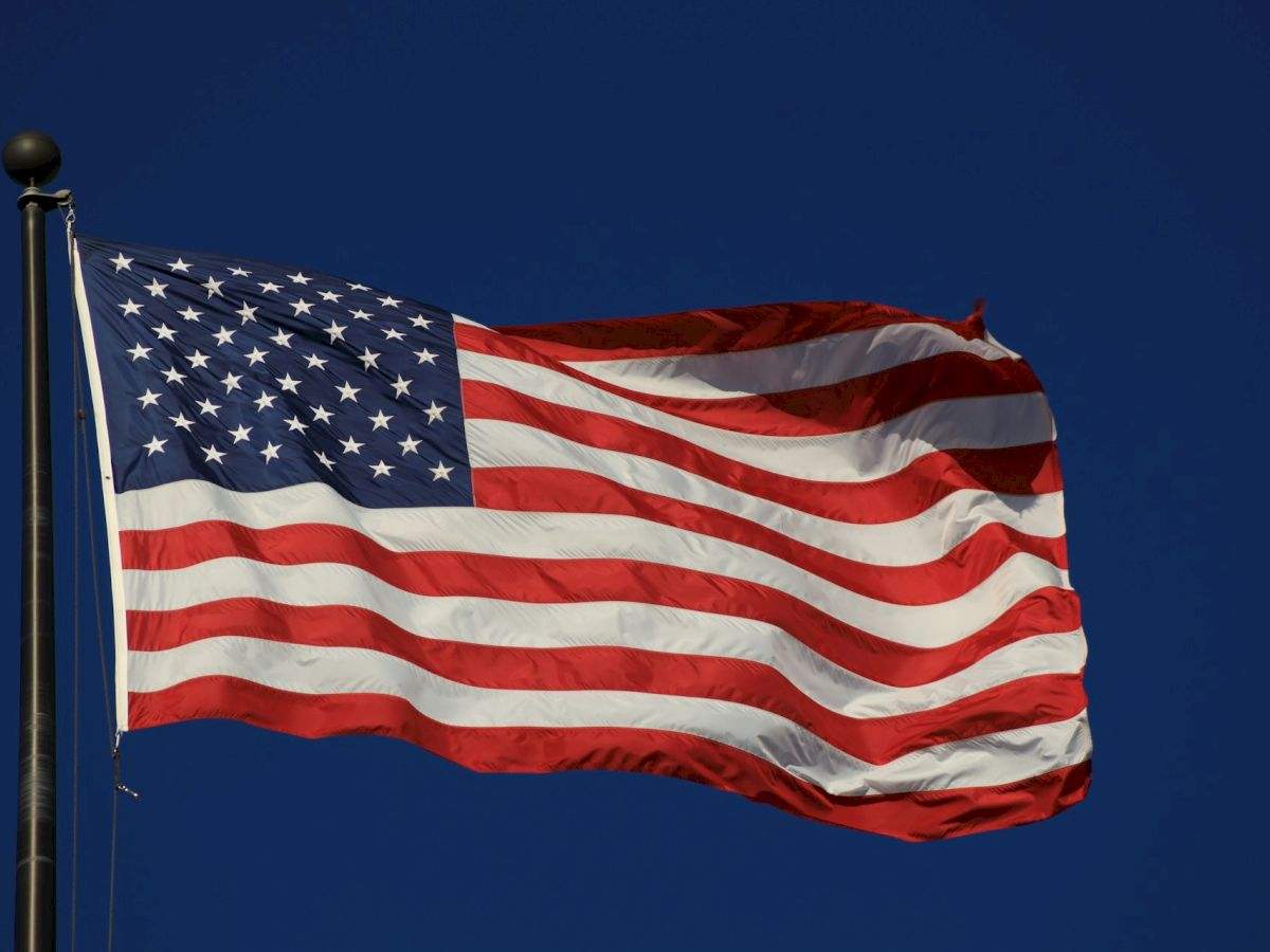 The image shows the flag of the United States, featuring stars and stripes, against a clear blue sky.