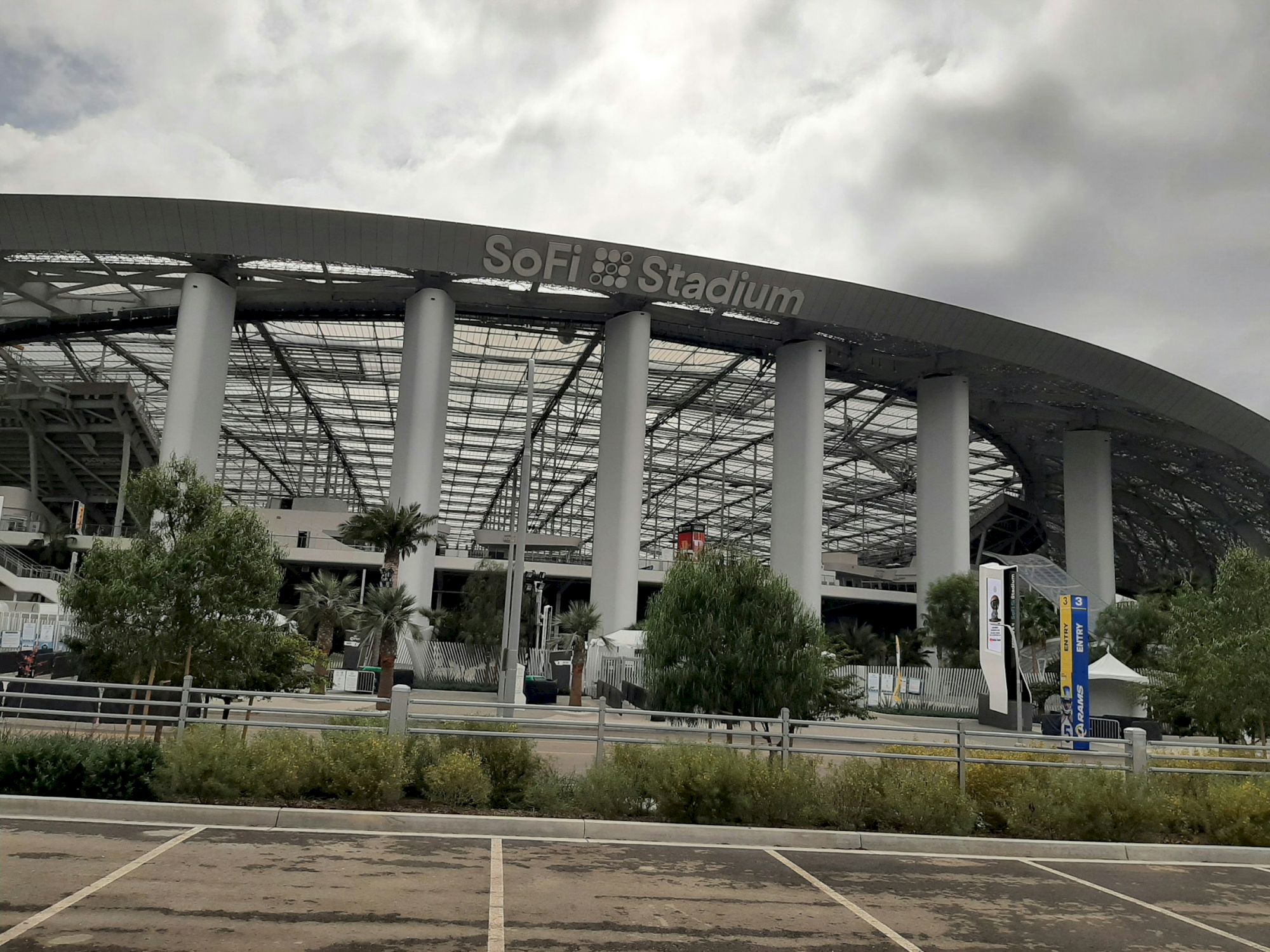 The image shows the exterior of SoFi Stadium, featuring its distinctive architecture and surrounding greenery under a cloudy sky.