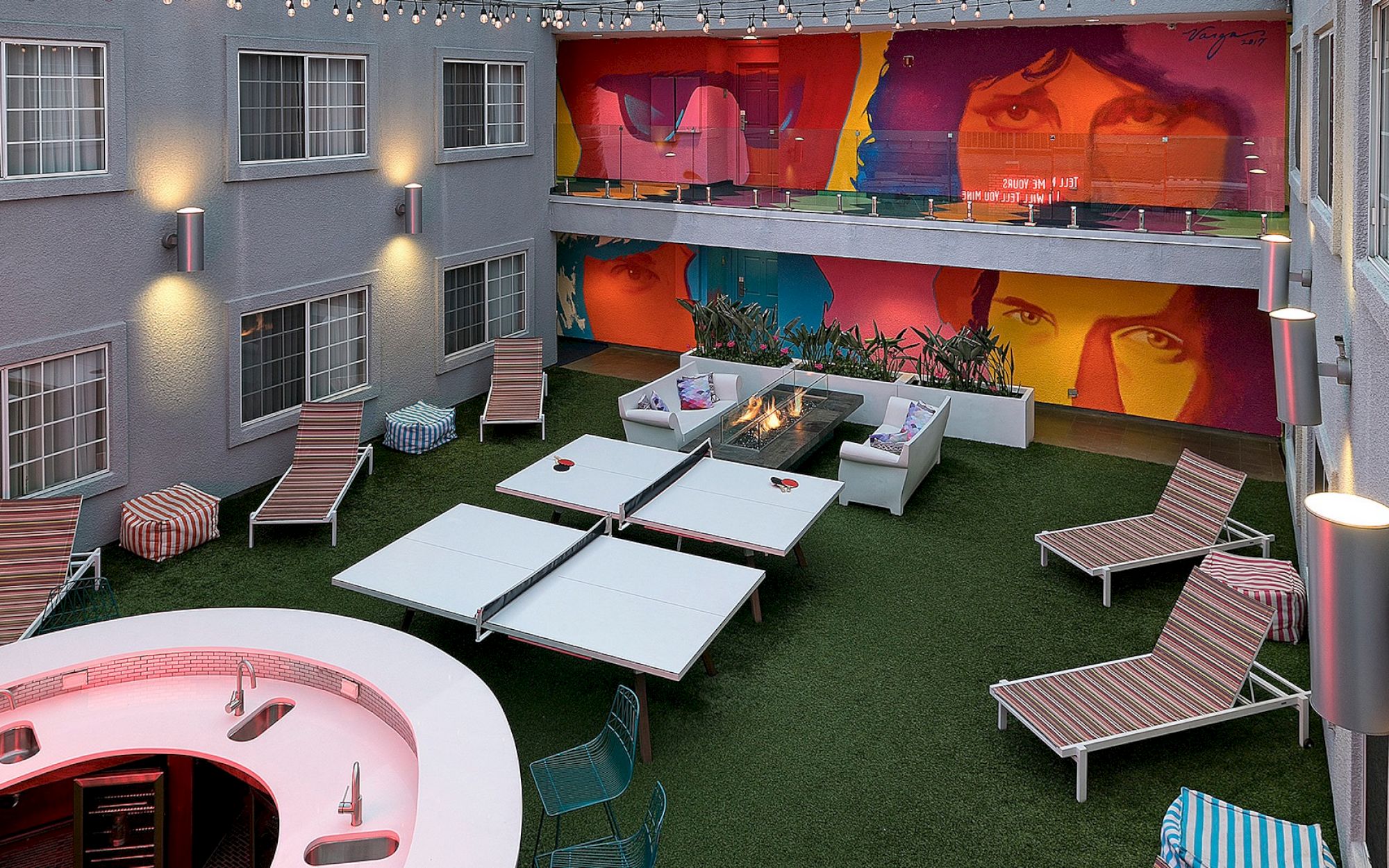 Outdoor lounge area with colorful murals, lounge chairs, tables, a fire pit, and string lights adorning the space for ambiance.