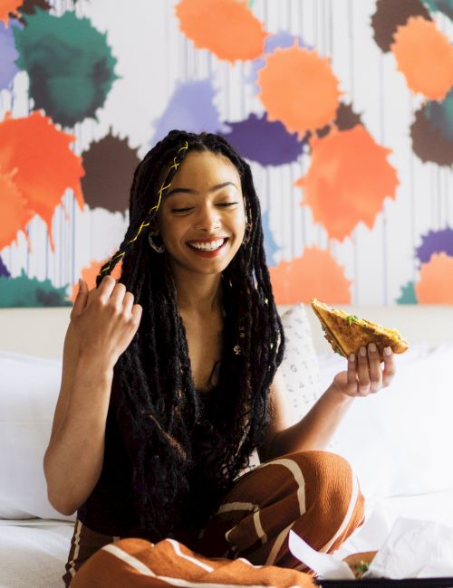 A person with long hair is sitting on a bed, smiling, holding a sandwich, with a colorful abstract wall behind them.