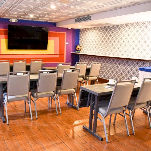 The image shows a small conference room with arranged chairs, tables, a large screen, patterned walls, and hardwood flooring.