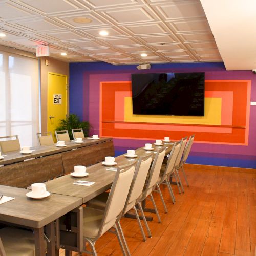 A conference room with a colorful wall design, large screen, tables, chairs, cups, and an "EXIT" sign.
