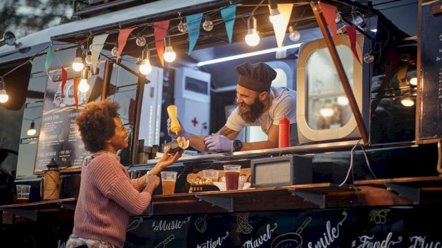 A food truck scene with a vendor in a chef's hat handing food to a customer, featuring colorful lights and decorative banners.