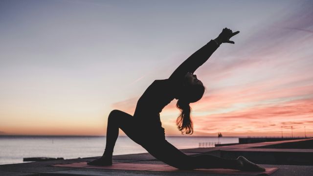 A person doing yoga in a lunge position at sunrise or sunset near the water, silhouetted against the colorful sky.