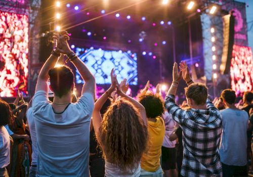 People are enjoying a live concert, clapping and cheering, with bright stage lights and a colorful backdrop in the background.