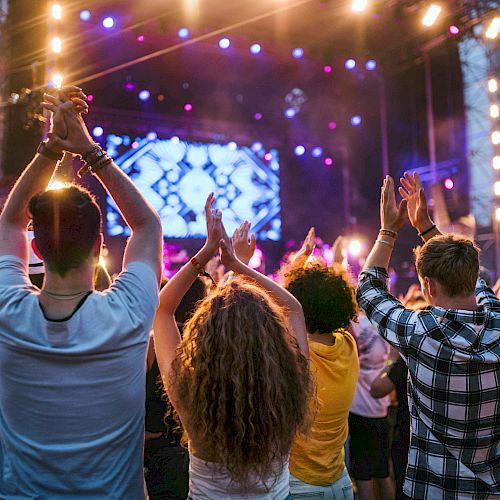 People are enjoying a live concert, clapping and cheering, with bright stage lights and a colorful backdrop in the background.