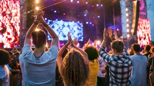 People are enjoying a live concert, clapping and cheering, with bright stage lights and a colorful backdrop in the background.