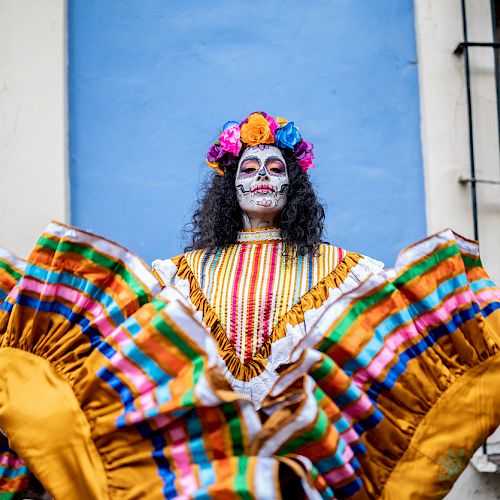 A person in colorful traditional attire and skull face paint, likely for Día de los Muertos, stands against a blue wall, holding up their skirt.