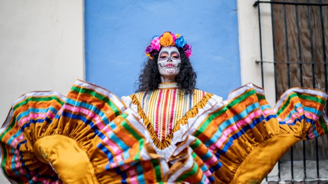A person in colorful traditional attire and skull face paint, likely for Día de los Muertos, stands against a blue wall, holding up their skirt.